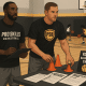 Two former pro basketball players set up a youth basketball camp in a gym with kids dribbling in the background and registration flyers on a table.