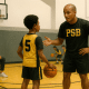 Coach instructing players while a parent watches respectfully during a youth basketball practice, in PSB brand colors.