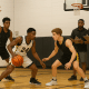 Four high school players compete during a basketball tryout as two coaches observe and take notes in a school gym with bright lighting and polished floors.