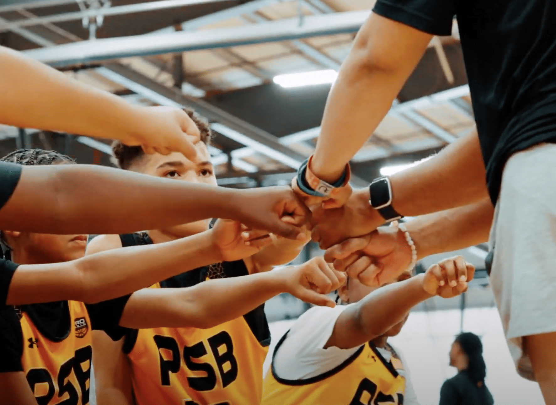 Youth basketball players in PSB yellow jerseys joining hands in team unity at the gym